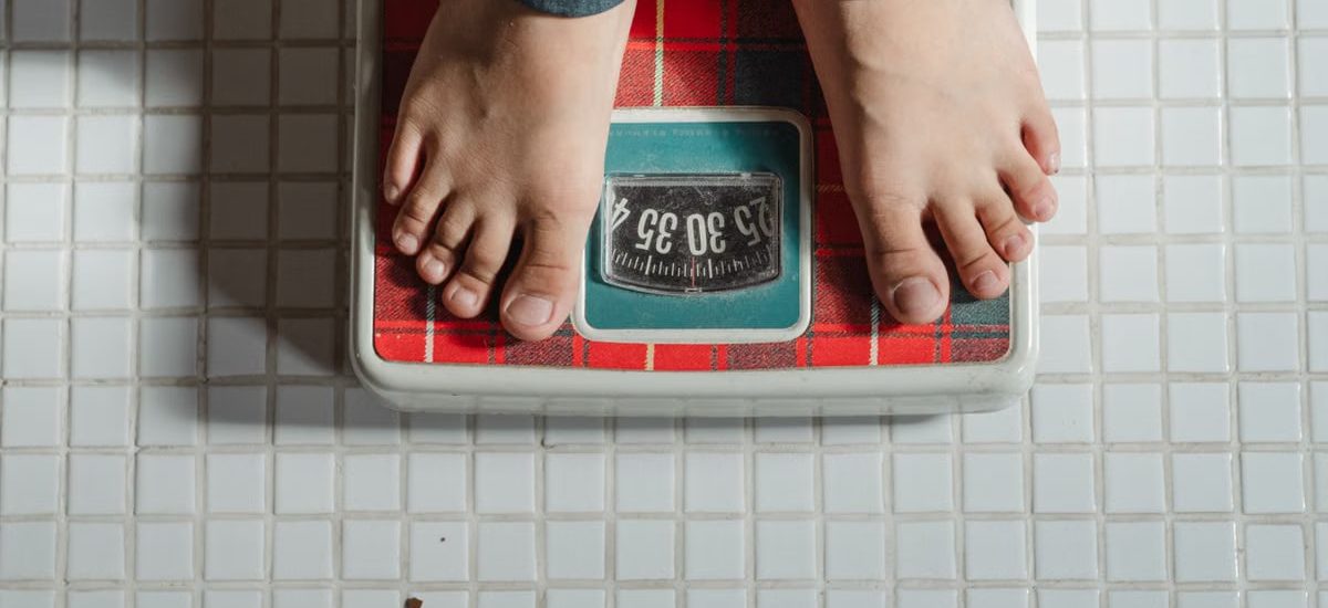 From above crop anonymous barefoot child in jeans standing on weigh scales on tiled floor of bathroom